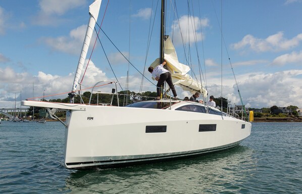 A white yacht with a cabin and a sail, docked at a marina with other boats and houses in the background.