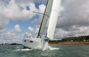 A sailboat with a white hull and sails, moving through a body of water with a rocky shoreline and trees in the background.