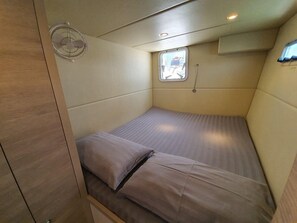 Una habitación pequeña con cama individual, ventana, ventilador y estante.