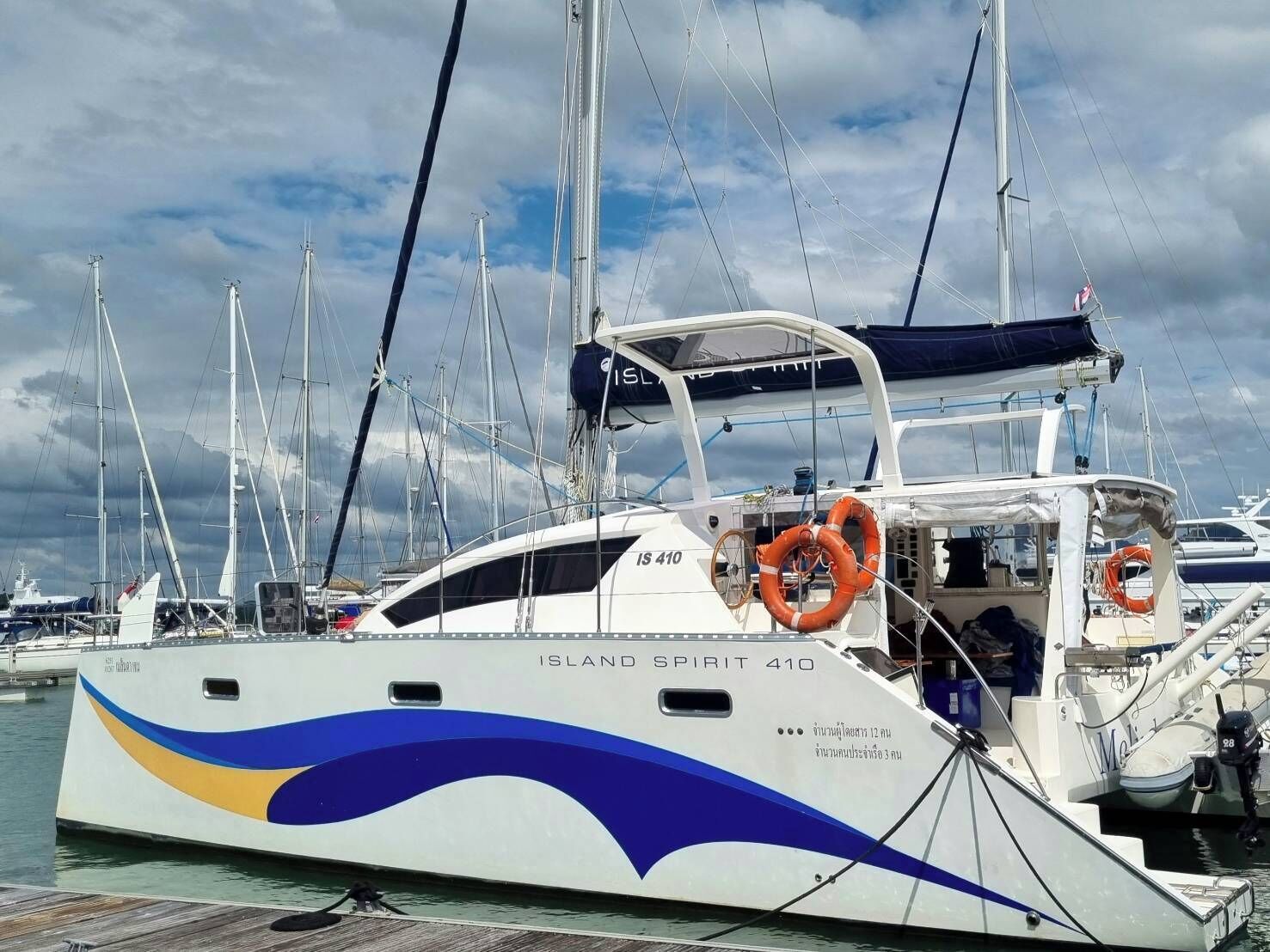 A yacht named 'Island Spirit 410' docked at a marina with multiple masts and other boats in the background.