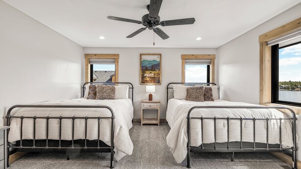 Quarto com duas camas de metal, um ventilador de teto, uma mesinha de cabeceira e janela com vista para as montanhas.