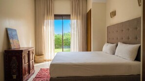 Ein Schlafzimmer mit einem großen Bett, einer Holzkommode und Blick auf Grün hinter einer Glastür.