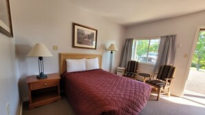 Una habitación de hotel con una cama grande, dos lámparas de noche, una mesita, una silla y una ventana con cortinas.