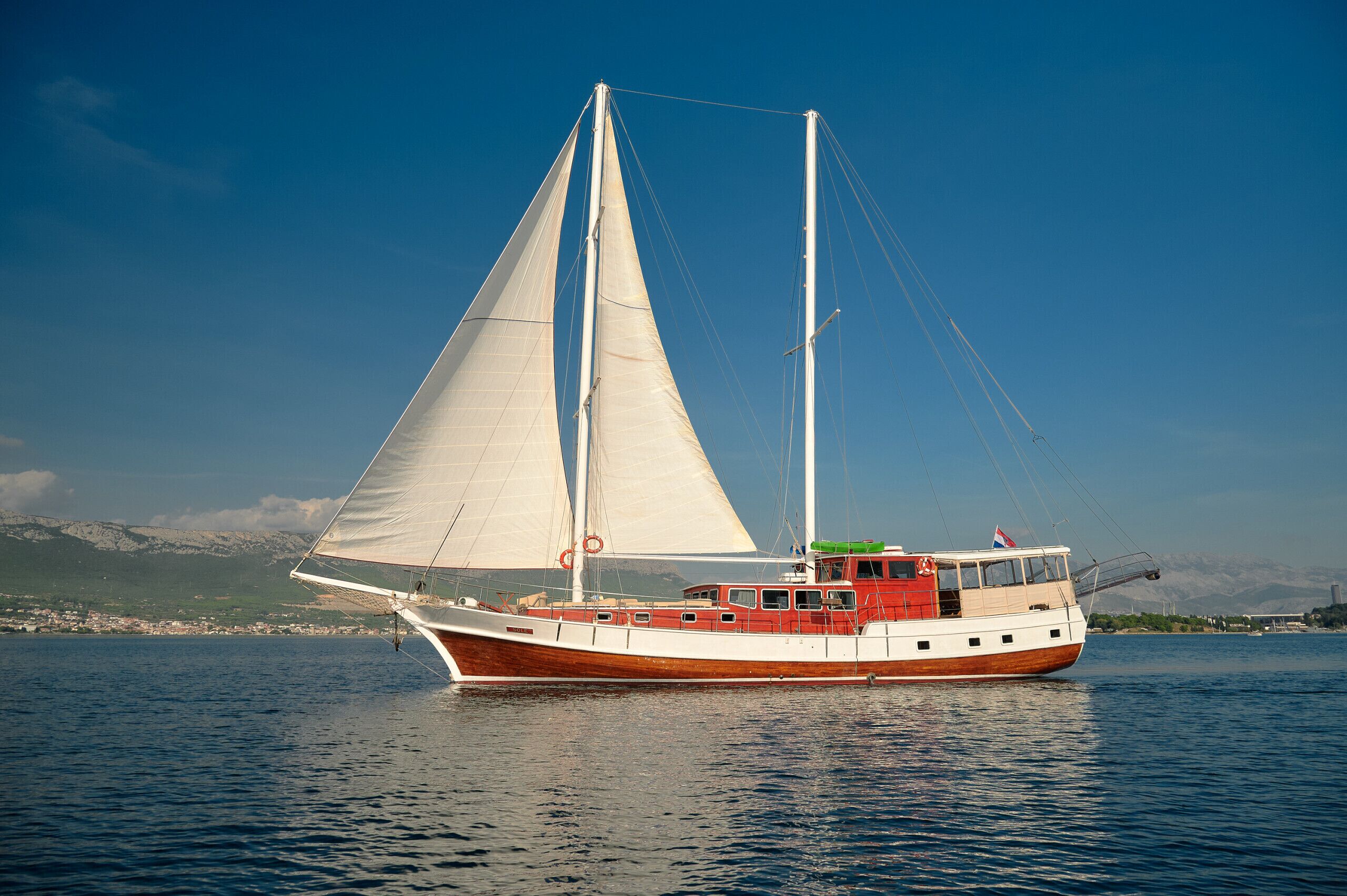 A boat with a cabin and sails, floating on water with a mountainous backdrop.