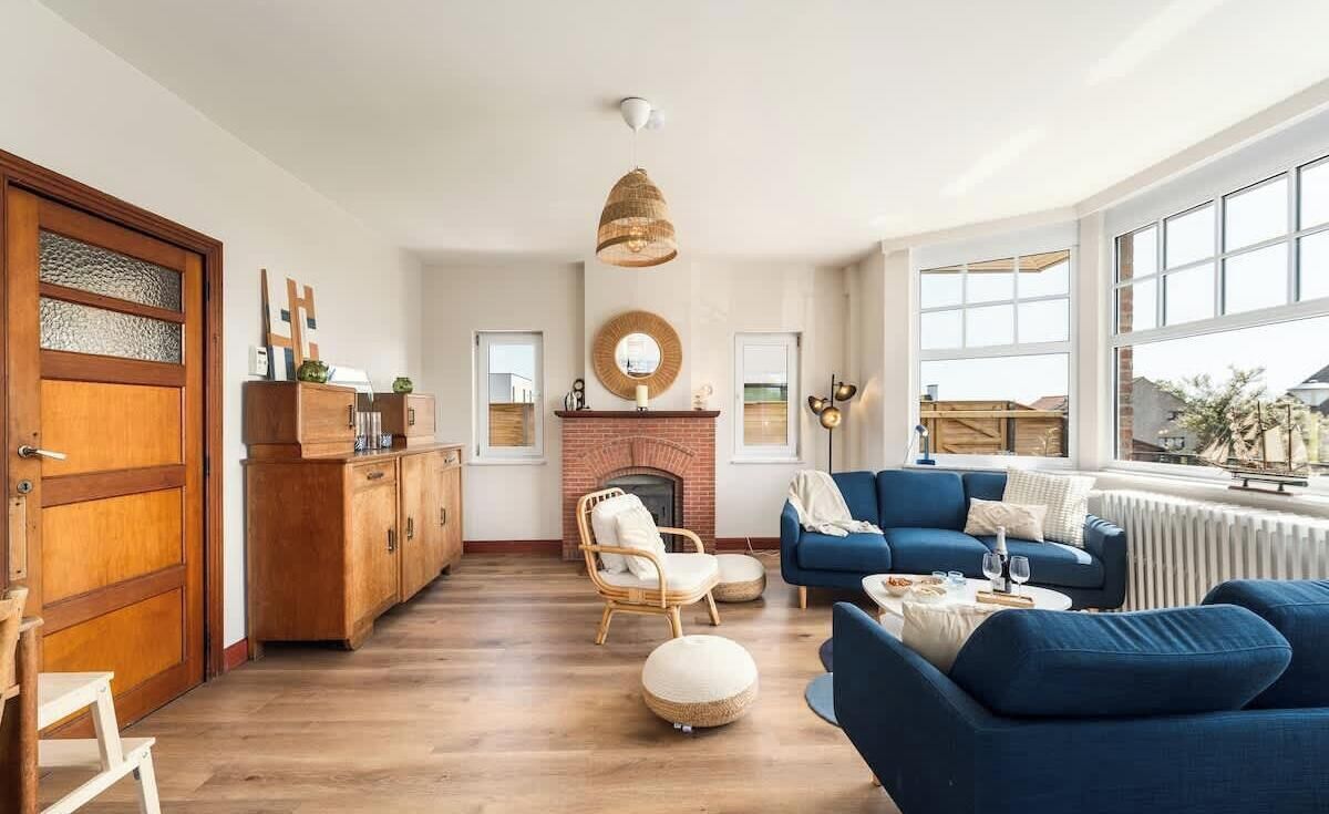 A spacious living room with a fireplace, blue sofas, a wooden cabinet, and a large window.