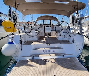 Dek van een boot met de naam 'Iustitia' op de boeg, stuurwiel en een gele reddingsboei.