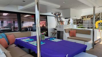 Interior de barco com mesa posta para refeição, assentos acolchoados e vista através de grandes janelas.