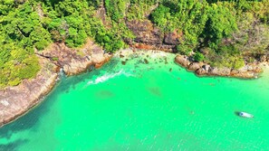 Luftaufnahme einer Küstenlandschaft mit klarem türkisblauem Wasser, felsigem Gelände und dichter Vegetation.