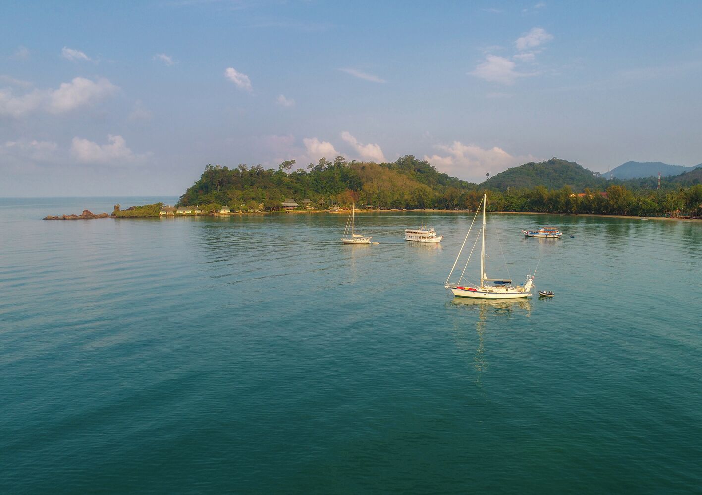 Una bahía tranquila con varios veleros anclados, una pequeña isla con edificios al fondo, y un cielo despejado.