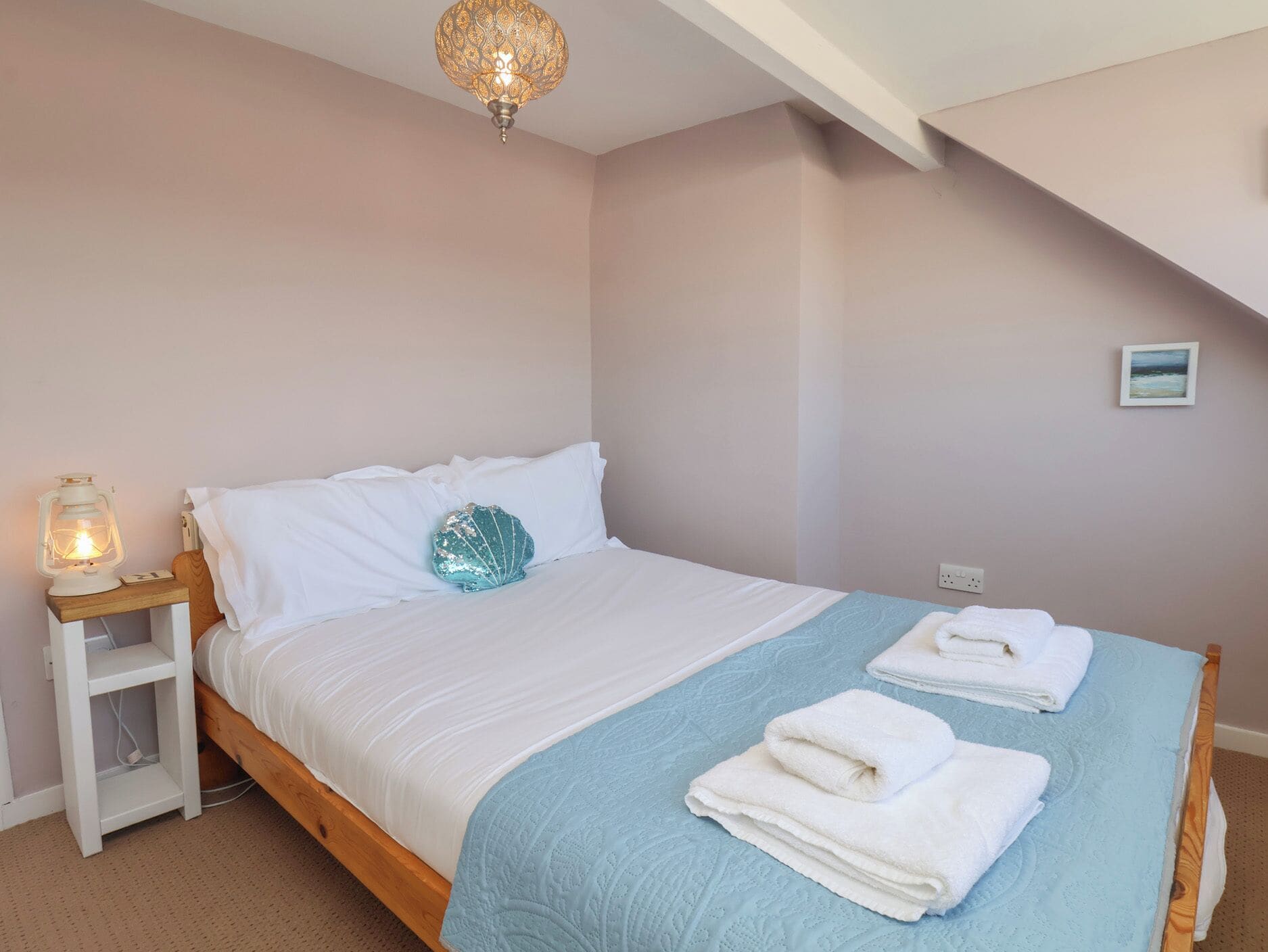Un dormitorio con una cama, una mesita de noche con un farol, un cojín decorativo con forma de caracola y un cuadro en la pared.