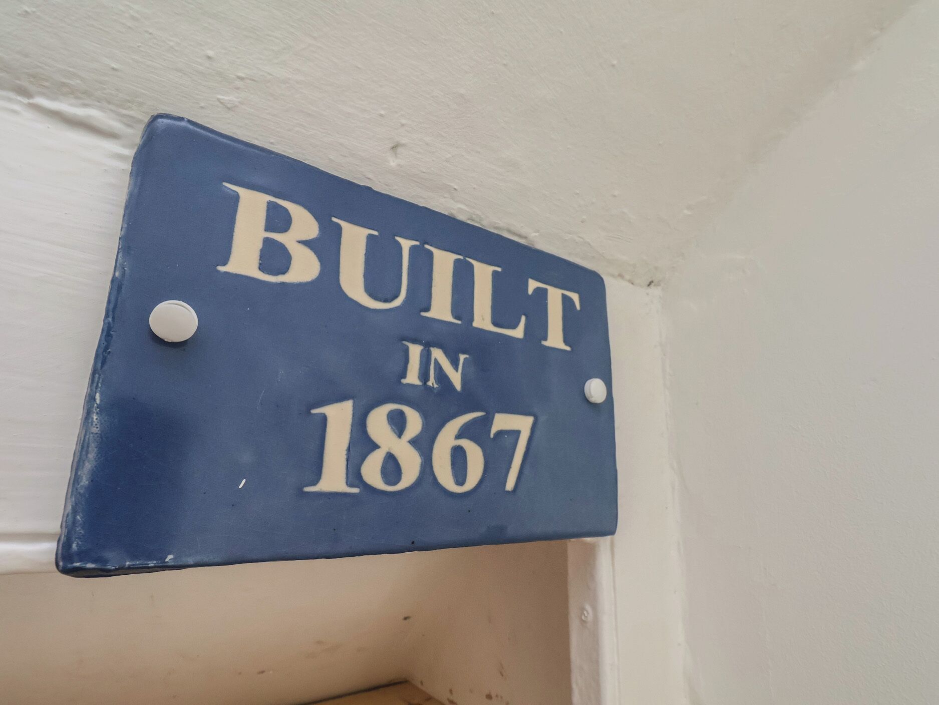 Une plaque bleue avec des lettres blanches indiquant « CONSTRUIT EN 1867 » est fixée à un mur blanc.