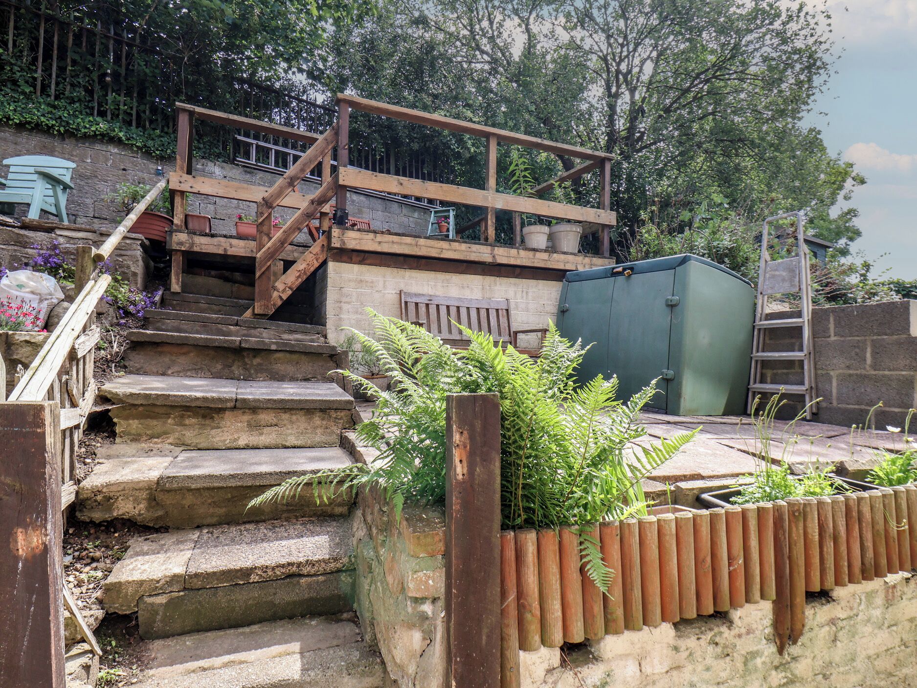 A wooden staircase leading to a deck with a railing, surrounded by greenery and a green shed.