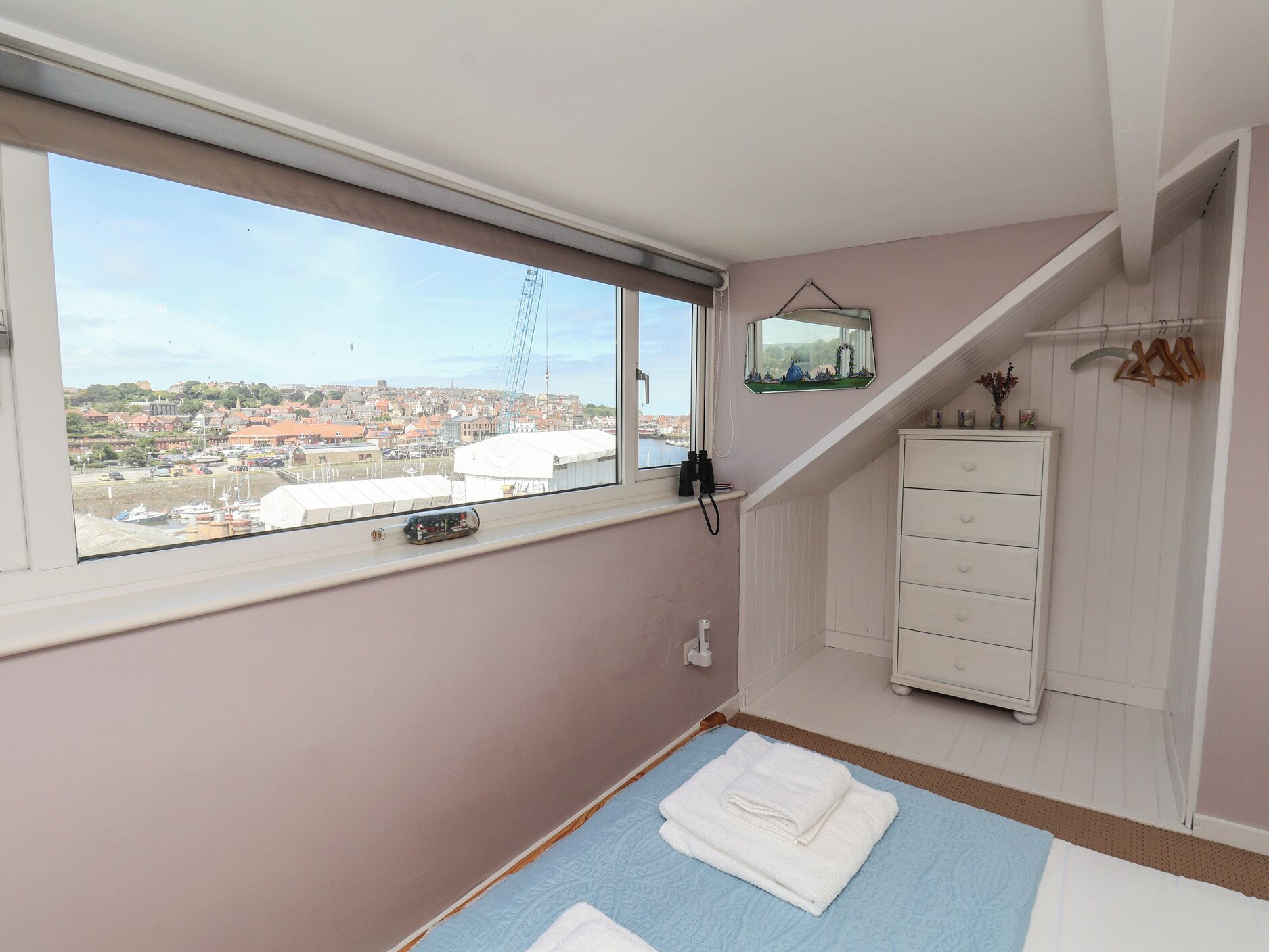 A bedroom with a large window, a bed with a blue and white bedspread, a white dresser, and a small mirror on the wall.