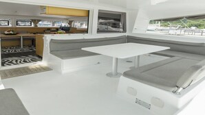 Interior de barco con comedor, cocina y zona de estar con mesa y cojines.