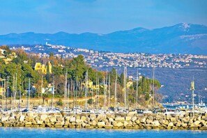A coastal town with a marina, rocky breakwater, and a backdrop of mountains.