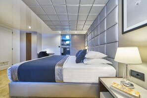 Ein modernes Hotelzimmer mit einem großen Bett, einem Nachttisch mit Lampe, einem an der Wand montierten Fernseher und einer Tür zu einem anderen Raum.