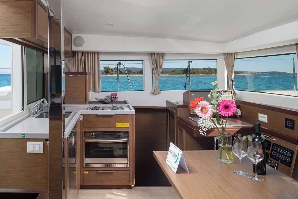 Uma cozinha compacta com fogão, forno e pia, um pequeno espaço bar com um vaso de flores e copos, e janelas amplas com vista para o mar.
