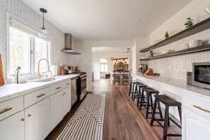 A modern kitchen with white cabinetry, a central island, bar stools, and a herringbone-patterned backsplash. - The Fireside: A Catskills Escape with Wide Open Spaces (Prattsville)