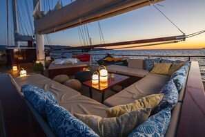 A yacht deck with a sofa area, cushions, a table with candles, and a view of the sea at sunset.
