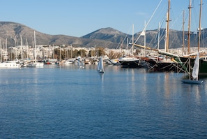 Un port de plaisance avec des voiliers, un voilier avec une personne à bord, et un paysage urbain avec des montagnes en arrière-plan.