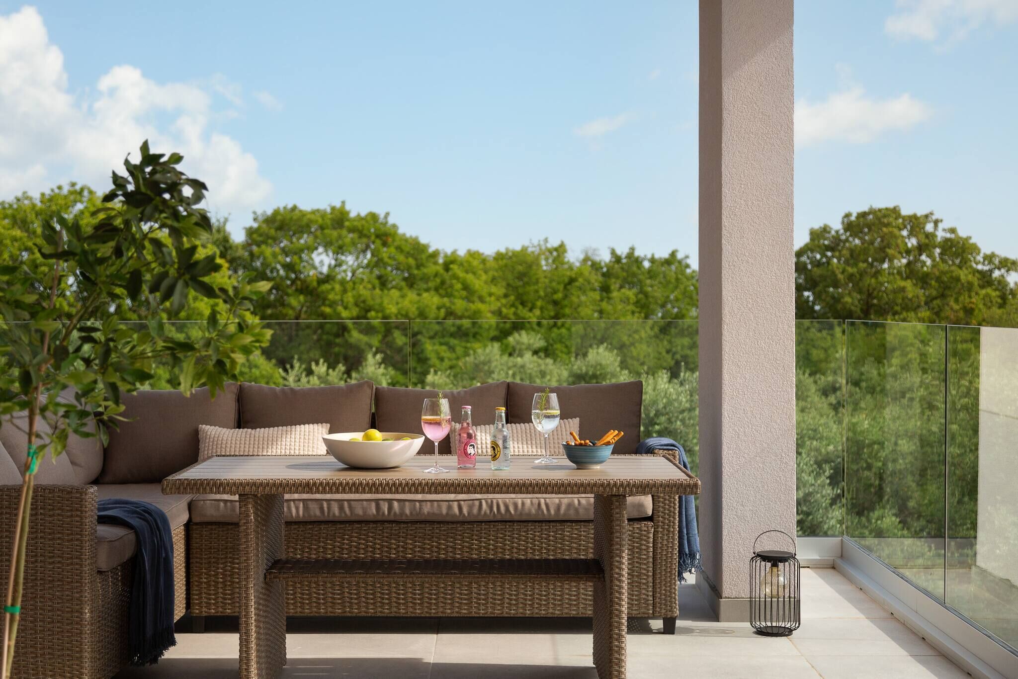 Un balcón con un sofá de mimbre, una mesa con bebidas y aperitivos, y vistas al verdor.
