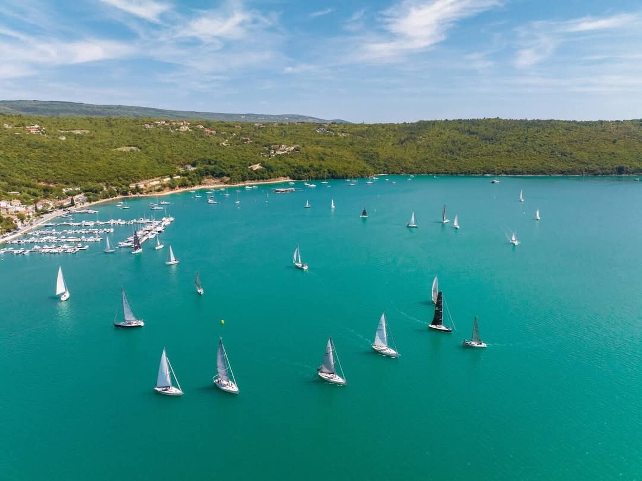 Ein Hafen mit mehreren Segelbooten, die in türkisblauem Wasser ankern, umgeben von grünen Hügeln und einem klaren Himmel.
