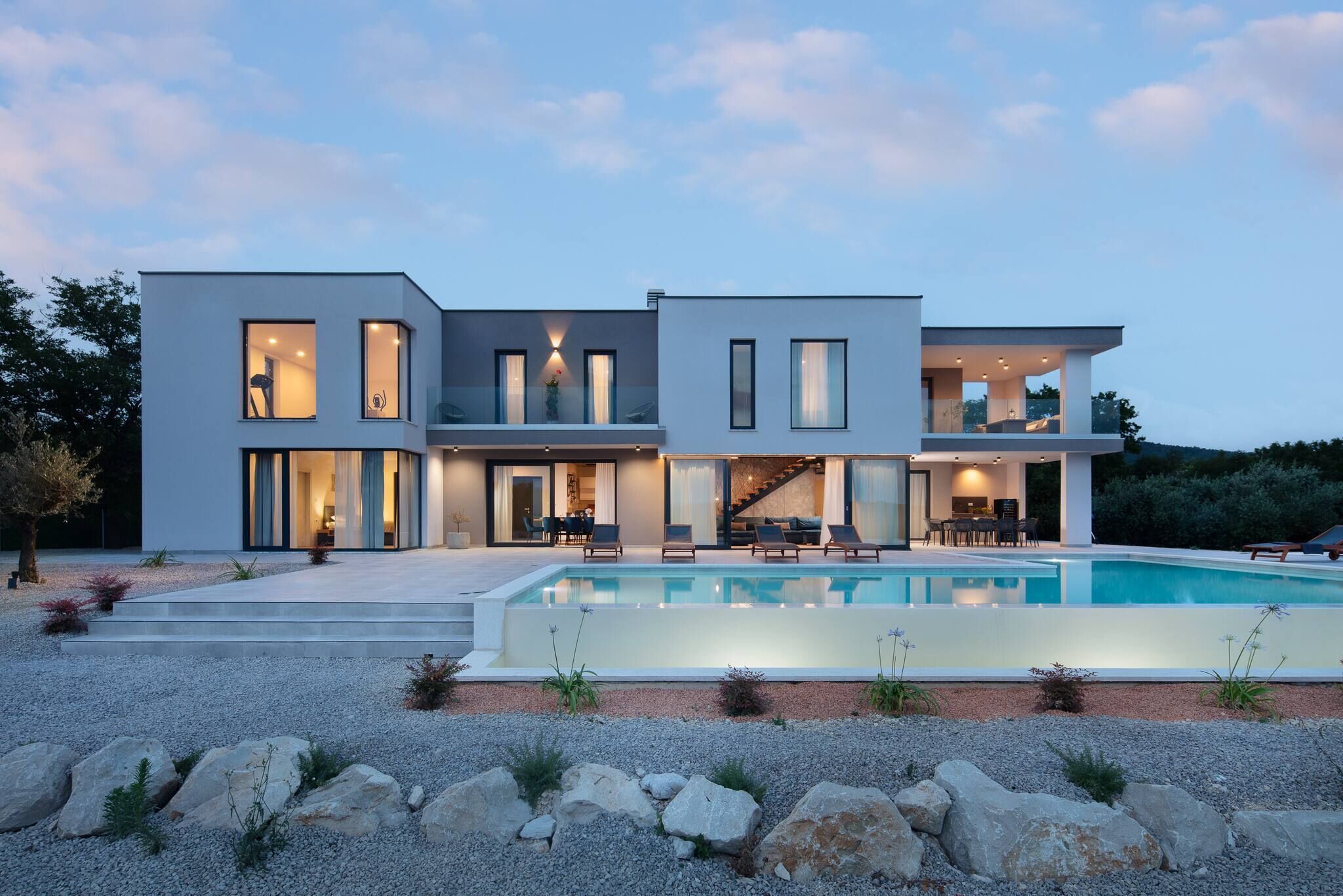 Ett modernt hus i två våningar med en swimmingpool, stora fönster och en väl upplyst interiör.