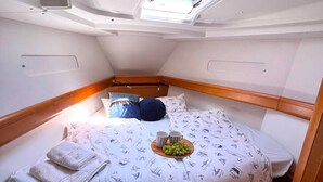En kompakt, veloplyst hytte med en seng med sengetæppe i maritim stil, et lille natbord med en kop og en bakke med frugt, samt et ovenlys.