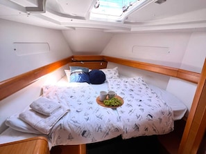 A compact, well-lit cabin with a bed featuring a nautical-themed bedspread, a small bedside table with a cup and a plate of fruit, and a skylight above.