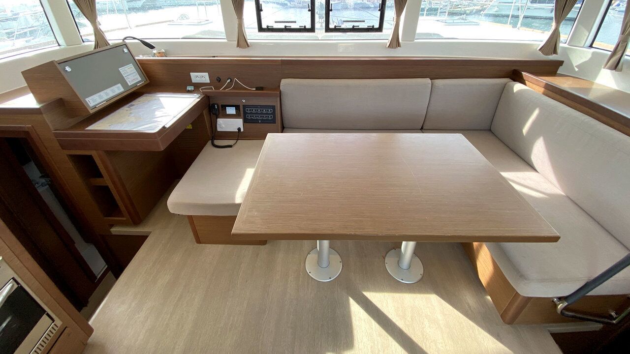 Un intérieur compact et bien éclairé, avec une table en bois et des sièges rembourrés, un panneau de commande et une fenêtre donnant sur le port de plaisance.