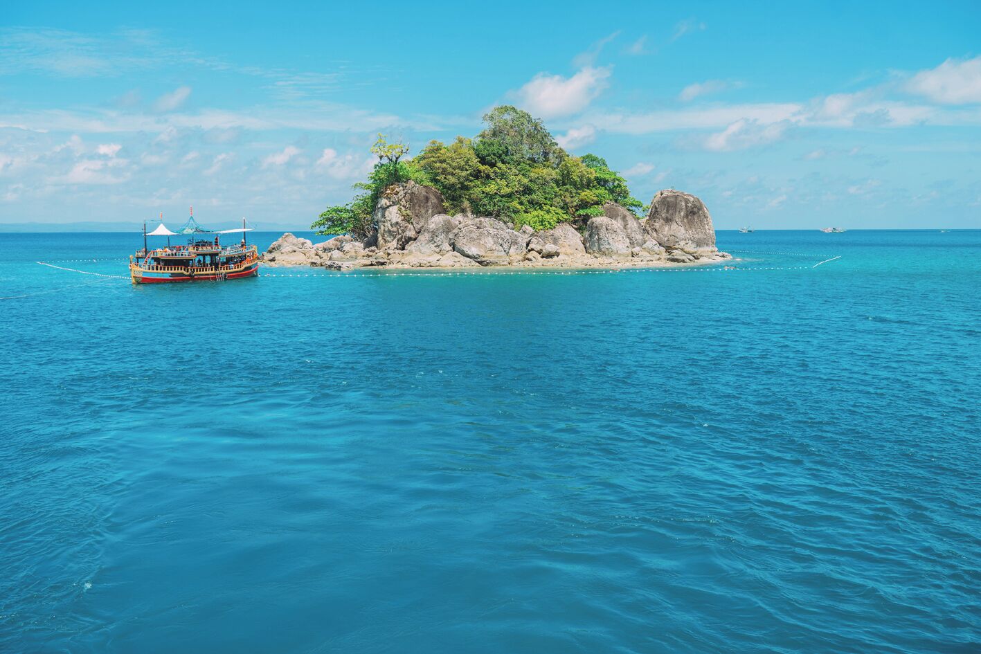 Un bateau sur une eau turquoise et limpide, une petite île rocheuse parsemée d’arbres et un ciel dégagé.