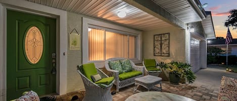 Un patio techado con mobiliario de mimbre, una puerta verde y una vista de la casa al fondo.