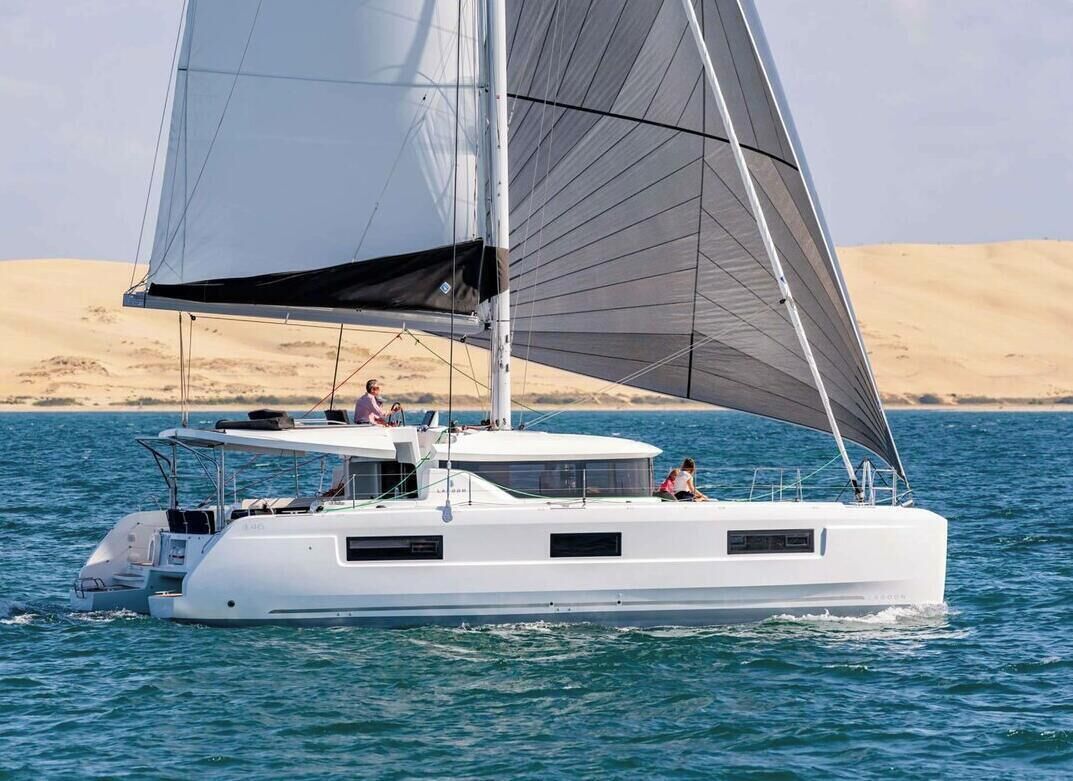 Un catamaran blanc à mât unique et à grande voile, naviguant sur des eaux calmes, avec une plage de sable en arrière-plan.