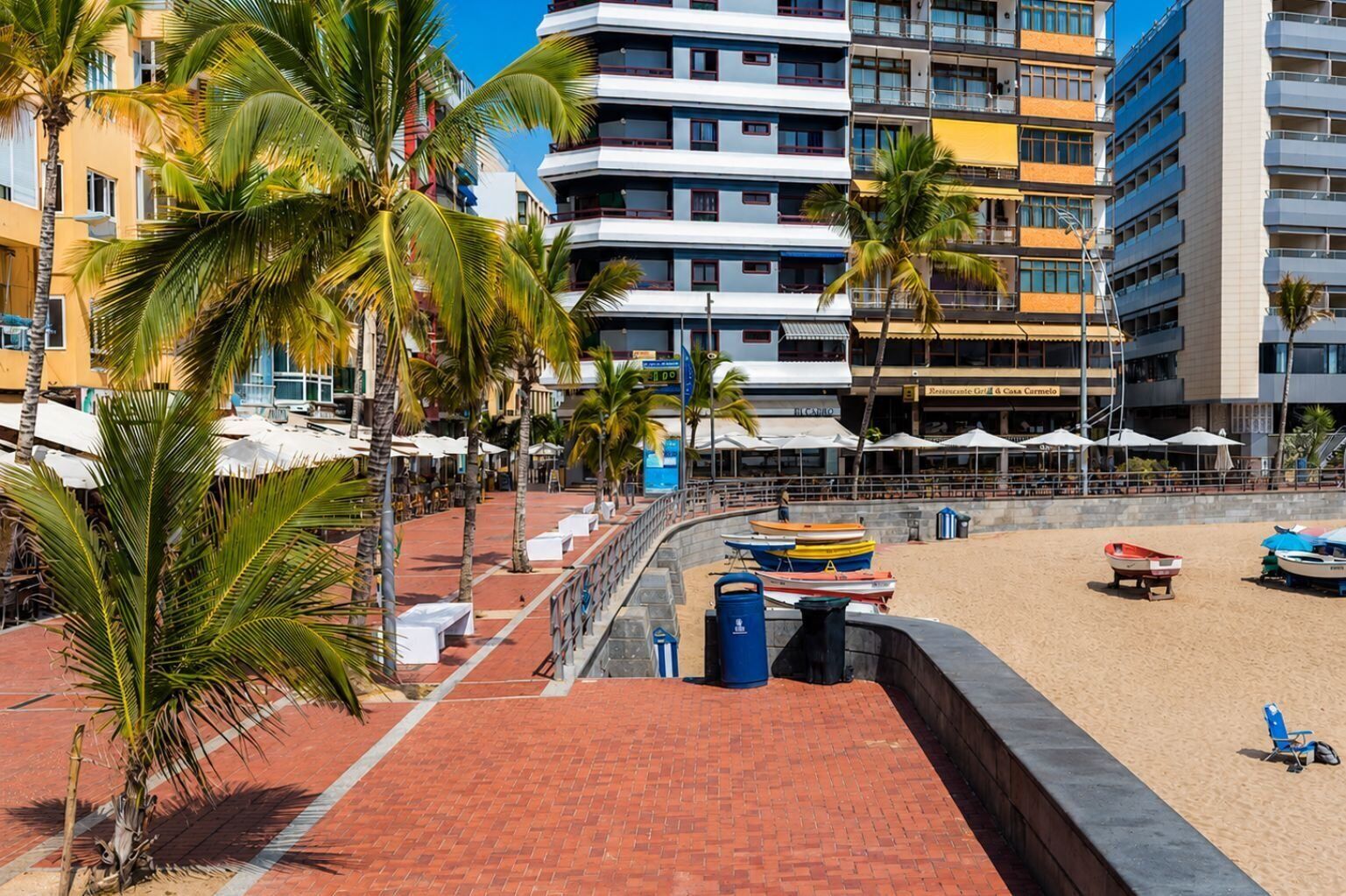 A beachfront hotel with a brick walkway, palm trees, and a sandy beach with boats.