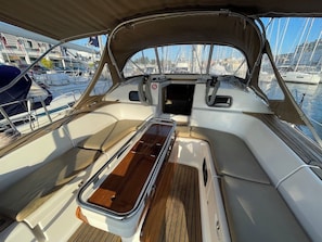 Het dek van een boot met een houten vloer, beklede zitplaatsen en uitzicht op andere boten en gebouwen.