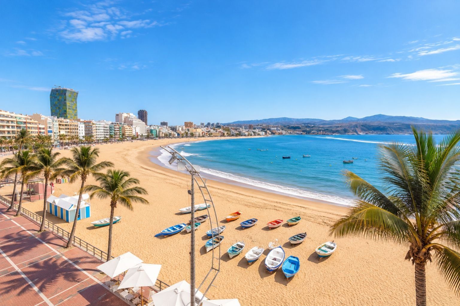 A beach with rows of boats, palm trees, and a cityscape in the background.