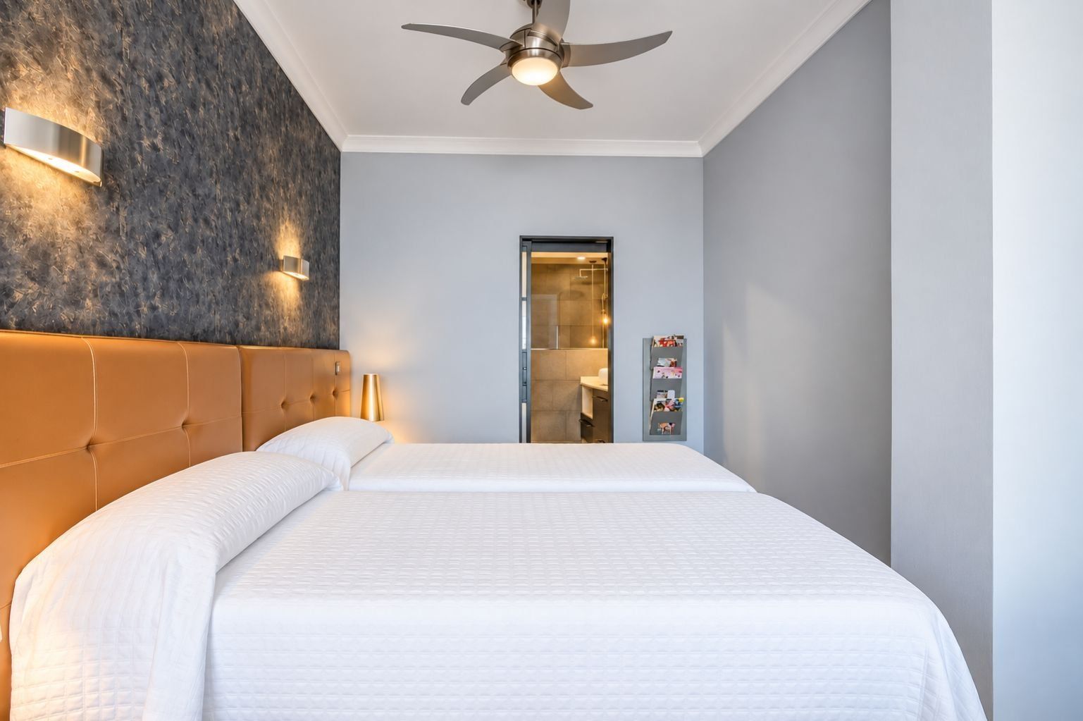 A hotel room with a large bed, a ceiling fan, a wall-mounted light, and a bathroom visible through a glass door.
