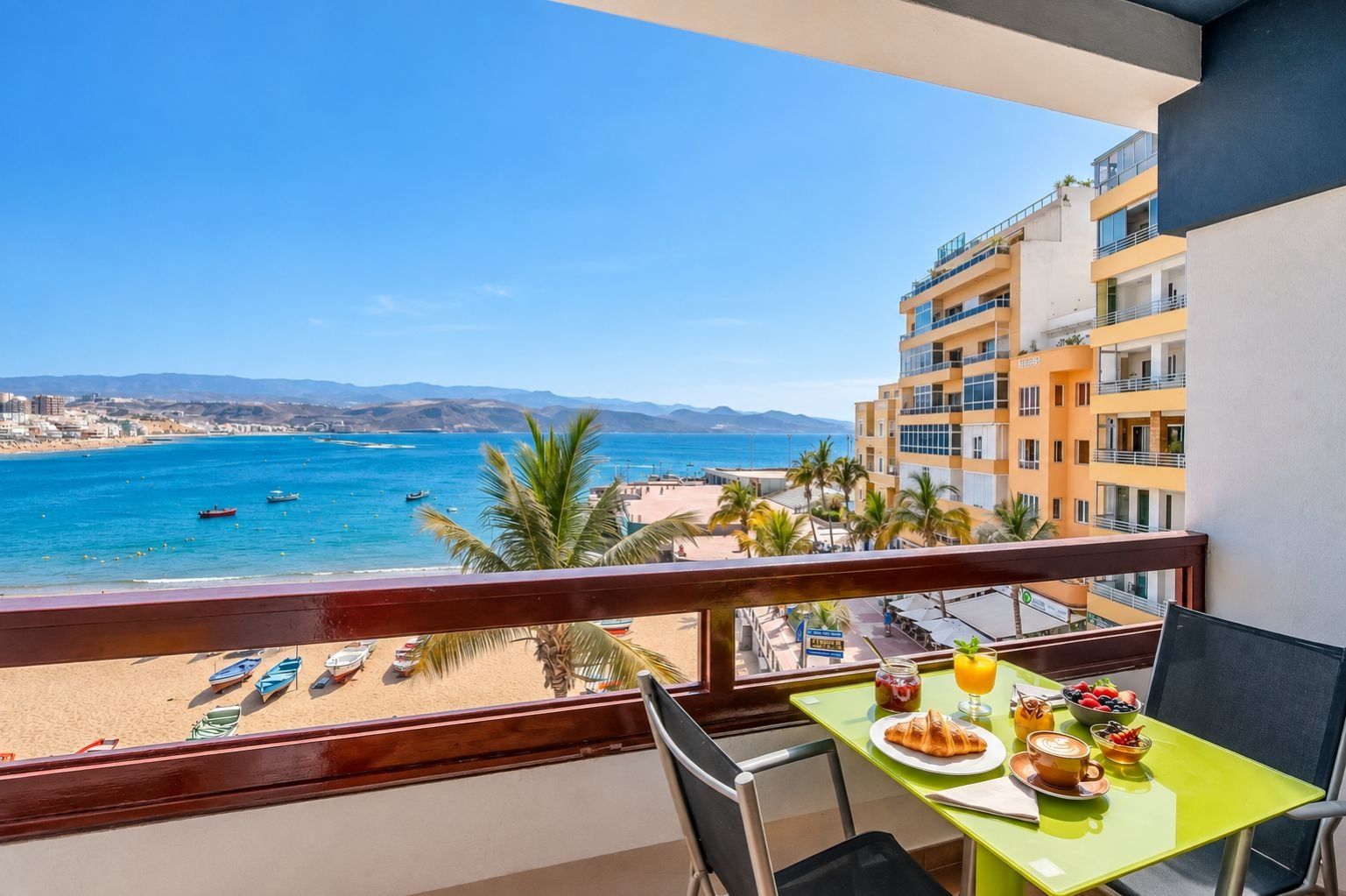Un balcón con mesa lista para el desayuno, con vista a una playa con barcos y palmeras.