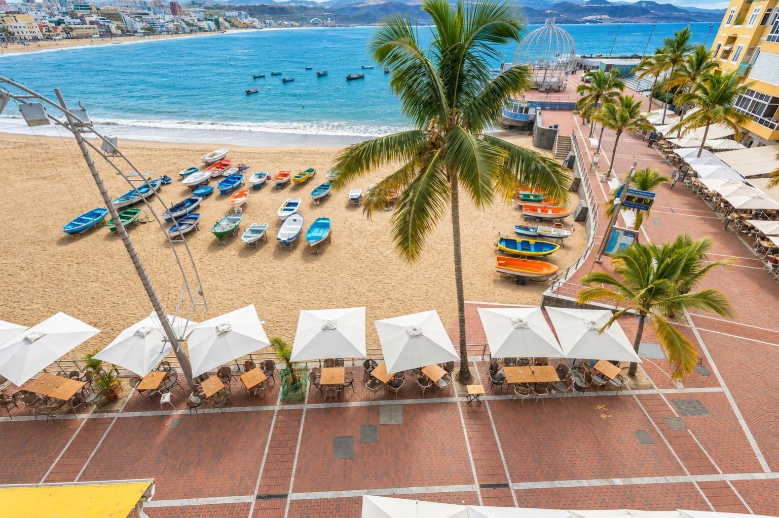 Vista de la playa con barcos, sombrillas y palmeras.