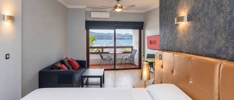 A hotel room with a bed, a sofa, a table, a chair, a balcony with a view, and a ceiling fan.