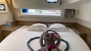 Ein ordentlich bezogenes Bett mit Handtüchern, ein Fenster mit Vorhängen und ein Nachttisch mit Wasserflaschen.