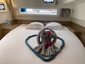 Ein ordentlich bezogenes Bett mit Handtüchern, ein Fenster mit Vorhängen und ein Nachttisch mit Wasserflaschen.
