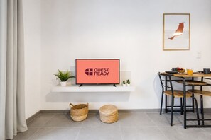 Ein modernes Zimmer mit einem Fernseher, der 'GUEST READY' anzeigt, einem kleinen Tisch mit Stühlen, einem Korb und einer Topfpflanze.
