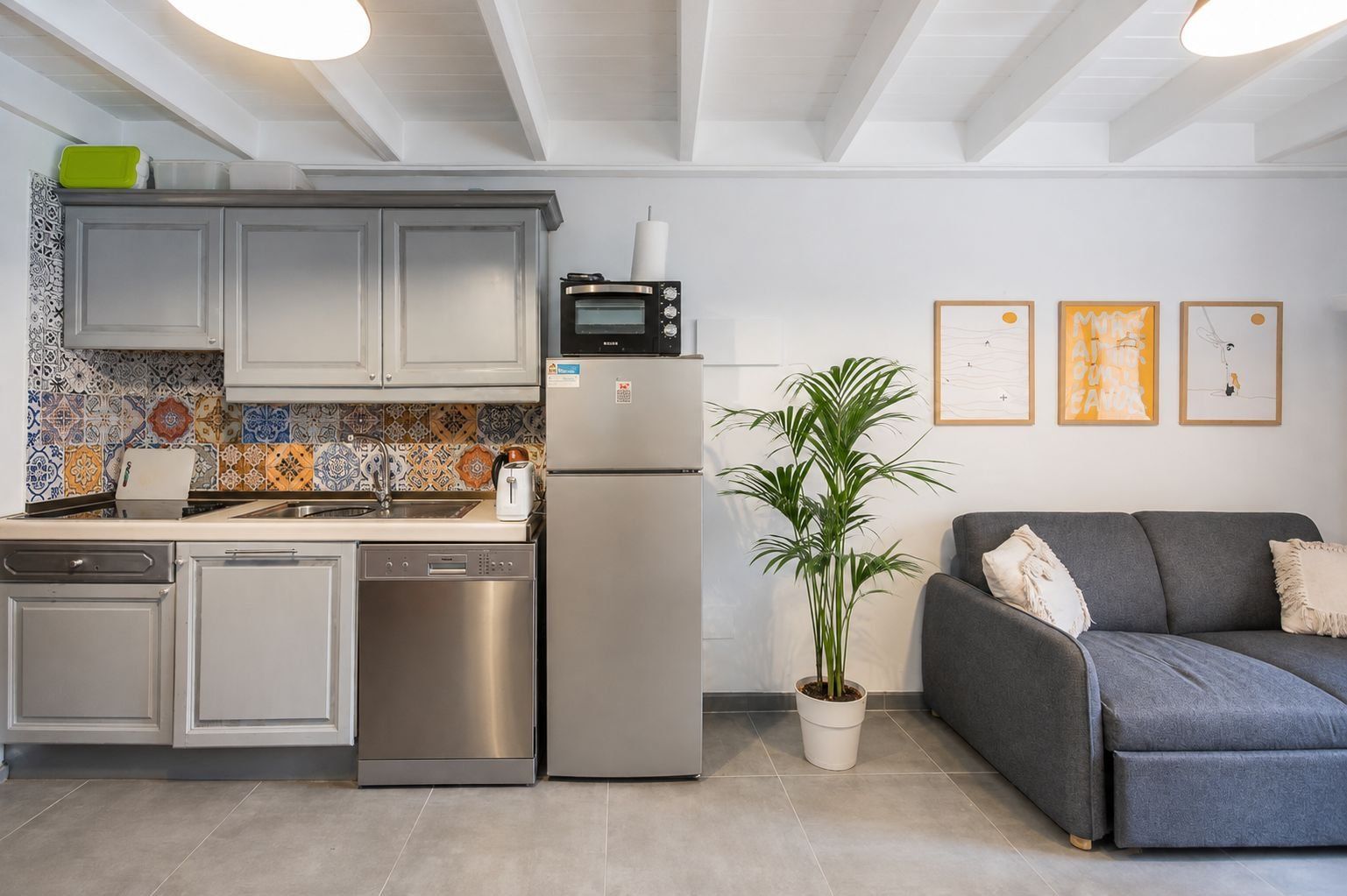 Una cocina moderna con horno integrado, refrigerador y lavavajillas. En la sala de estar, hay un sofá gris y una planta en maceta.