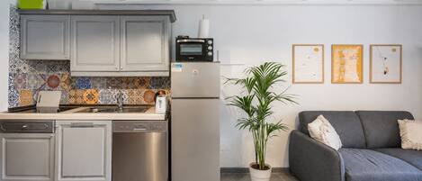 Una cocina moderna con horno integrado, refrigerador y lavavajillas. En la sala de estar, hay un sofá gris y una planta en maceta.