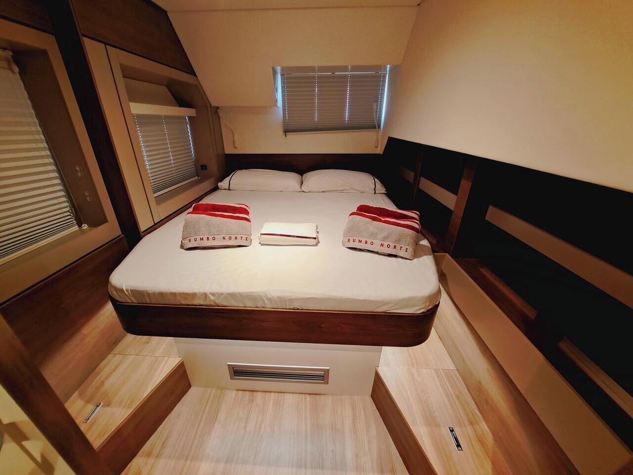 Een compact bed met hoofdeinde, een raam met zonwering, en een klein ventilatie rooster onder het bed.