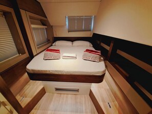 Ein kompaktes Bett mit Kopfteil, ein Fenster mit Jalousien und ein kleiner Lüftungsschlitz unter dem Bett.