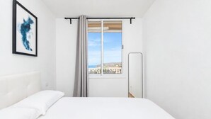 A hotel room with a bed, a window with a view of buildings, a framed abstract painting, and a mirror. - GuestReady - Balcon de Amadores Sea View (Puerto Rico)