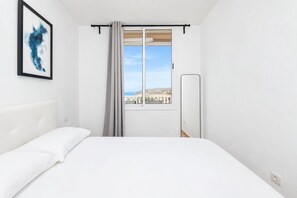 A hotel room with a bed, a window with a view of buildings, a framed abstract painting, and a mirror. - GuestReady - Balcon de Amadores Sea View (Puerto Rico)