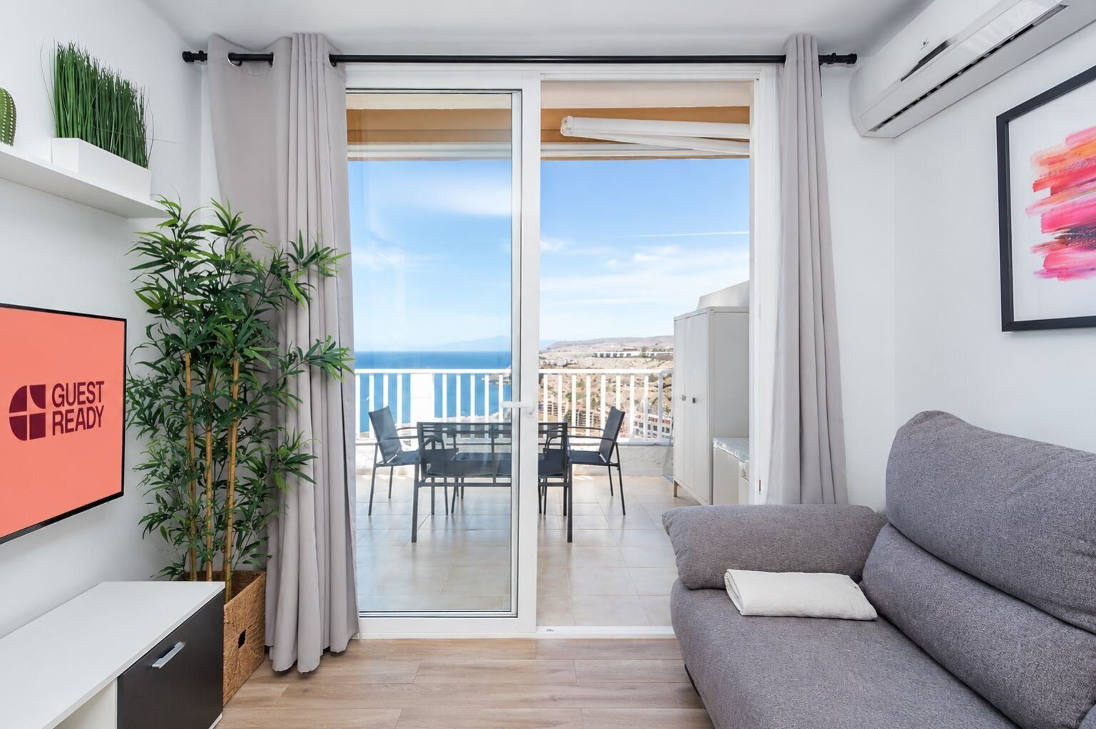 A modern living room with a grey sofa, a dining table with chairs, a large sliding glass door leading to a balcony with a view of the sea, and a 'GUEST READY' sign on the wall.